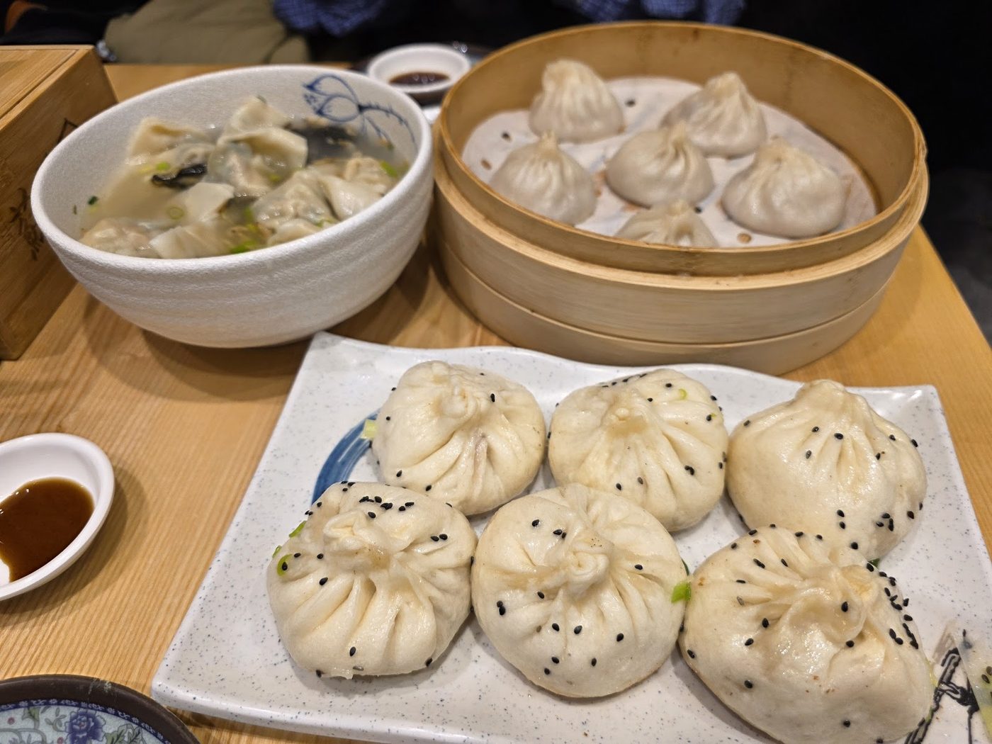 Bamboo steamer of xiao long bao beside a bowl of dumplings in broth and a plate of handmade buns