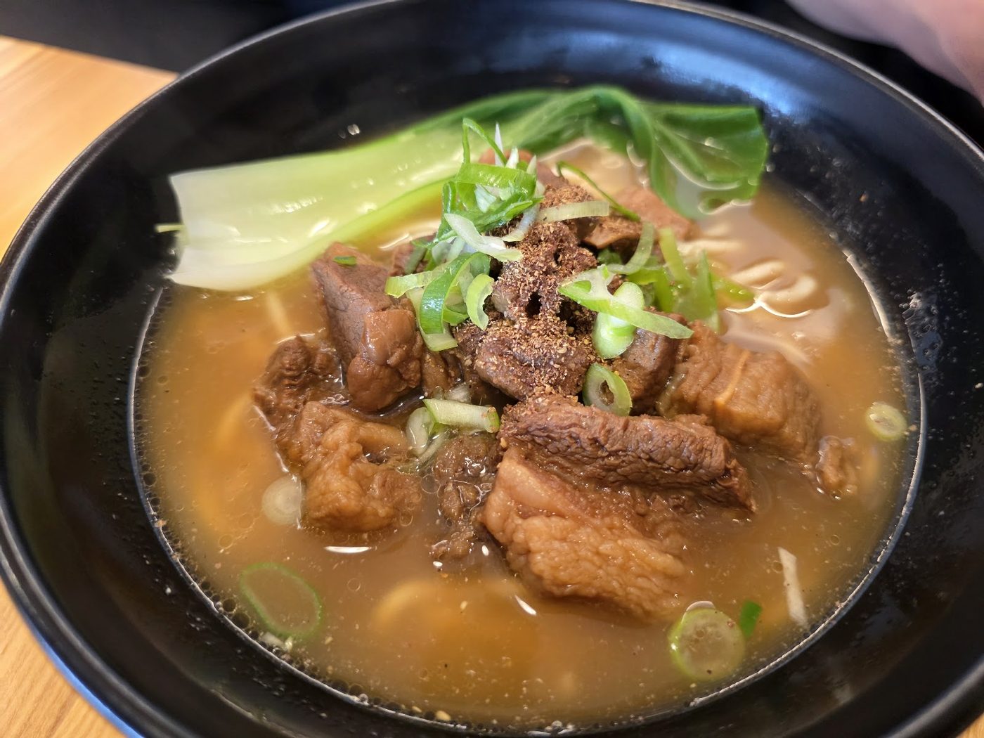 A deep black bowl of beef noodle soup with tender beef pieces, greens, spring onion and a rich broth