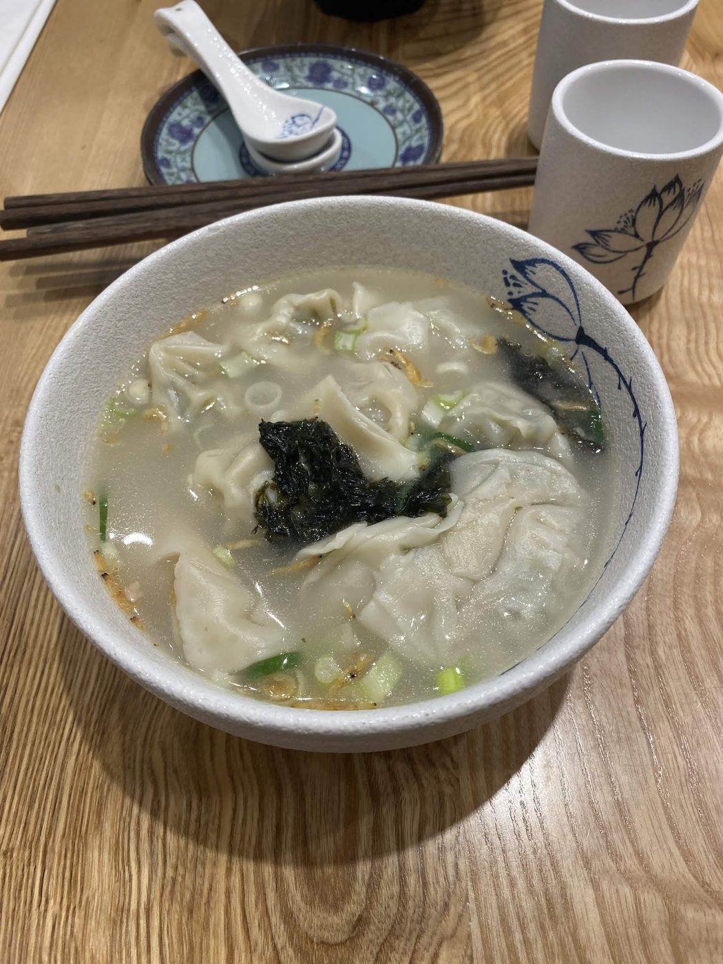 A bowl of dumplings in clear broth with seaweed and spring onion, with chopsticks and lotus-patterned cups behind