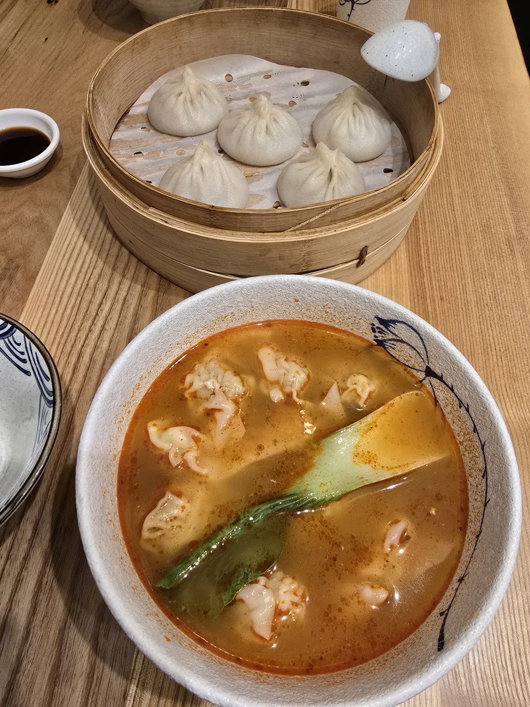 A bamboo steamer of soup dumplings beside a bowl of wontons in red broth and leafy greens